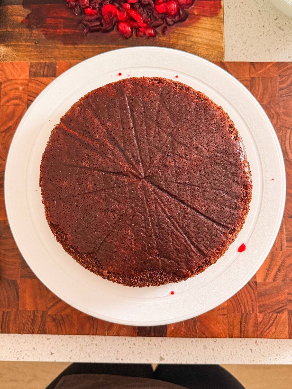 A round chocolate cake brushed with cherry syrup sits on a white plate on a wooden countertop. Red berry stains are visible on the cake edge and in the background.