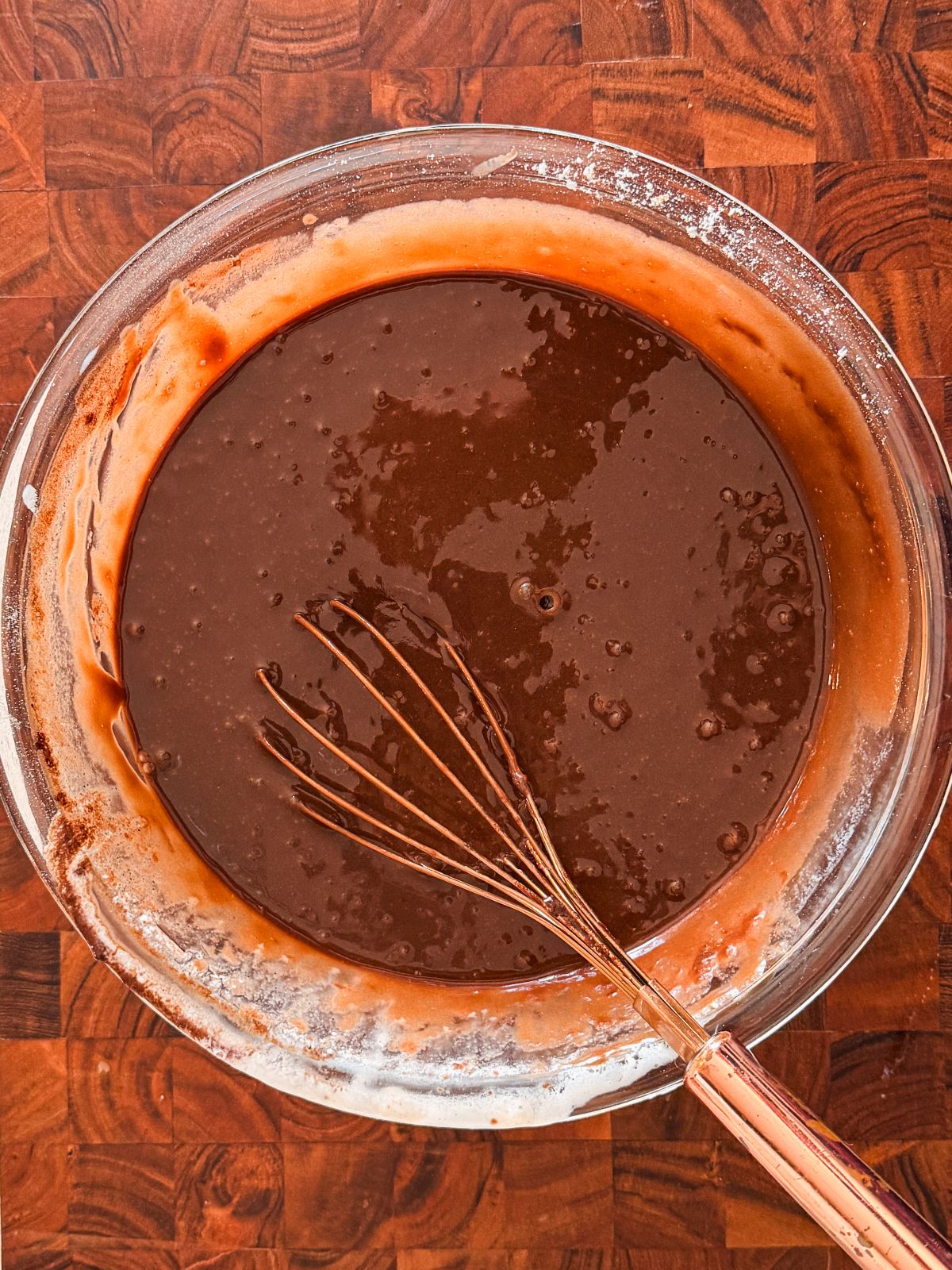 A glass bowl filled with chocolate cake batter sits on a wooden surface, with a metal whisk resting inside the bowl. Some batter is smeared along the sides of the bowl.
