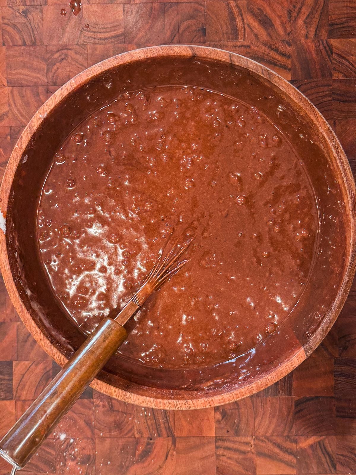 A wooden bowl filled with chocolate cake batter sits on a checkered wooden surface. A metal whisk with a wooden handle rests inside the bowl, partially covered in batter.