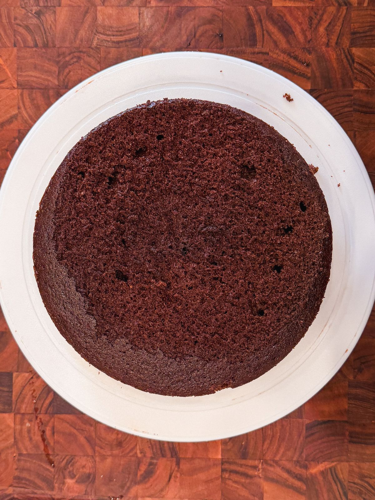 A round, unfrosted chocolate cake sits on a white plate atop a dark wooden surface. The cake has a smooth, slightly domed top with a few small holes.