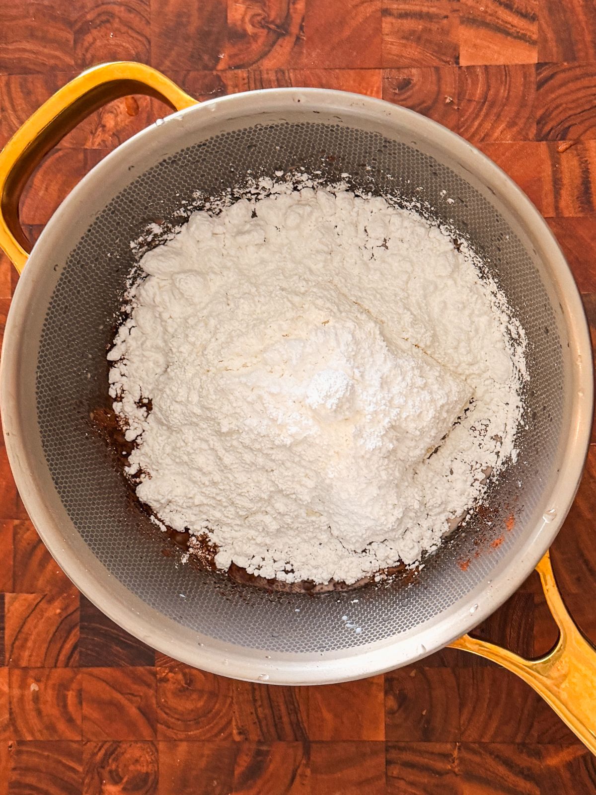 A metal mixing bowl with yellow handles contains a mound of white powdered sugar on top of a brown mixture, set on a wooden surface.