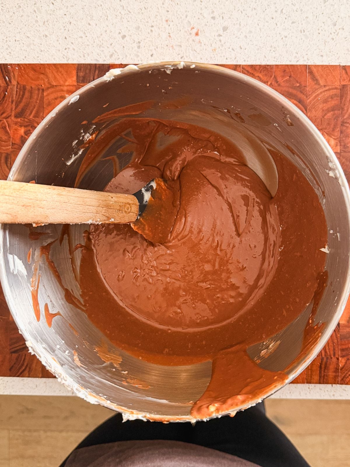 A metal mixing bowl filled with runny chocolate frosting and a spatula resting inside sits on a wooden cutting board on a countertop.