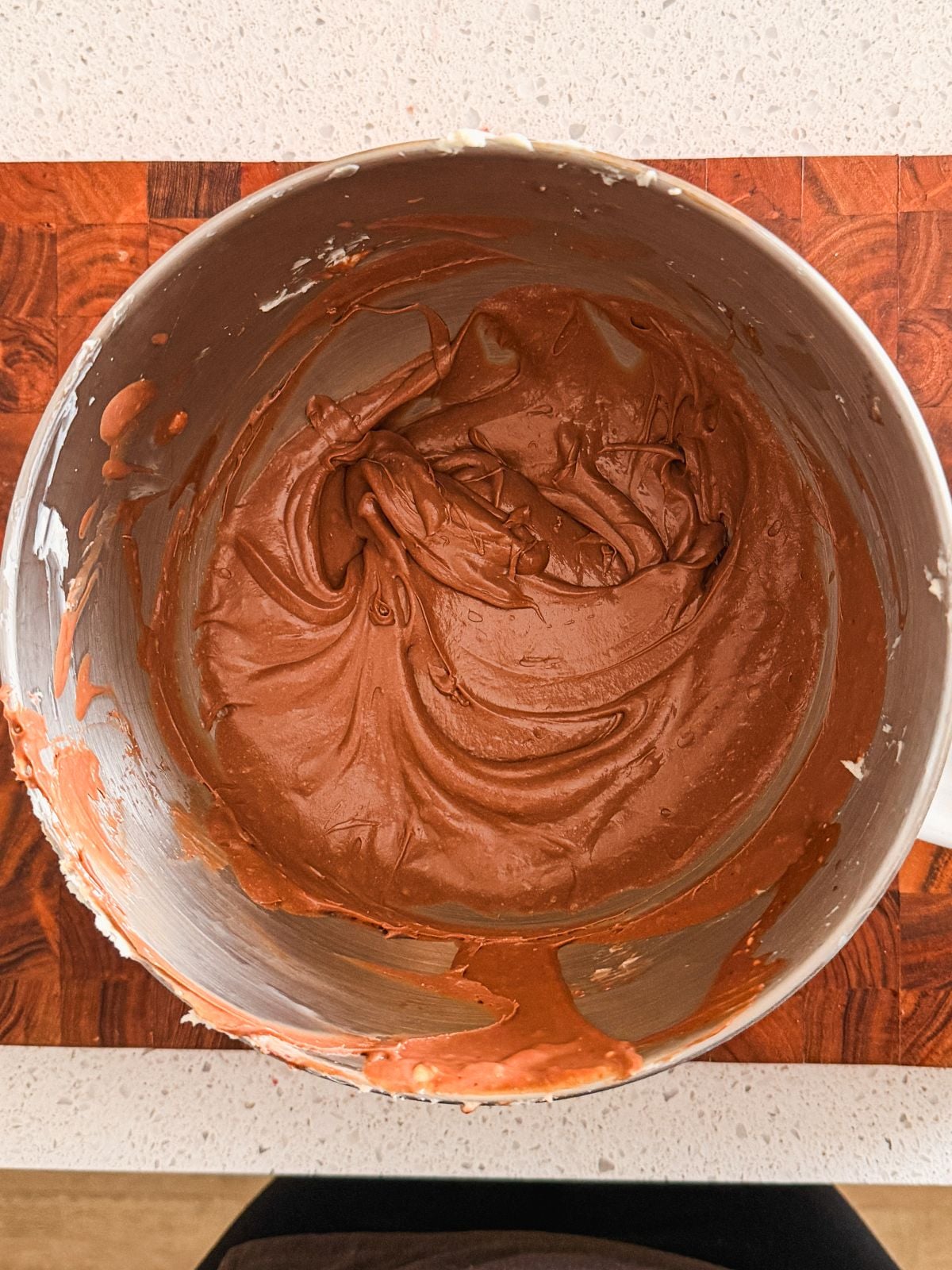 A mixing bowl filled with creamy chocolate frosting sits on a wooden surface. Batter is thick and swirled, with some smudges on the bowls edges. The countertop underneath is light-colored.