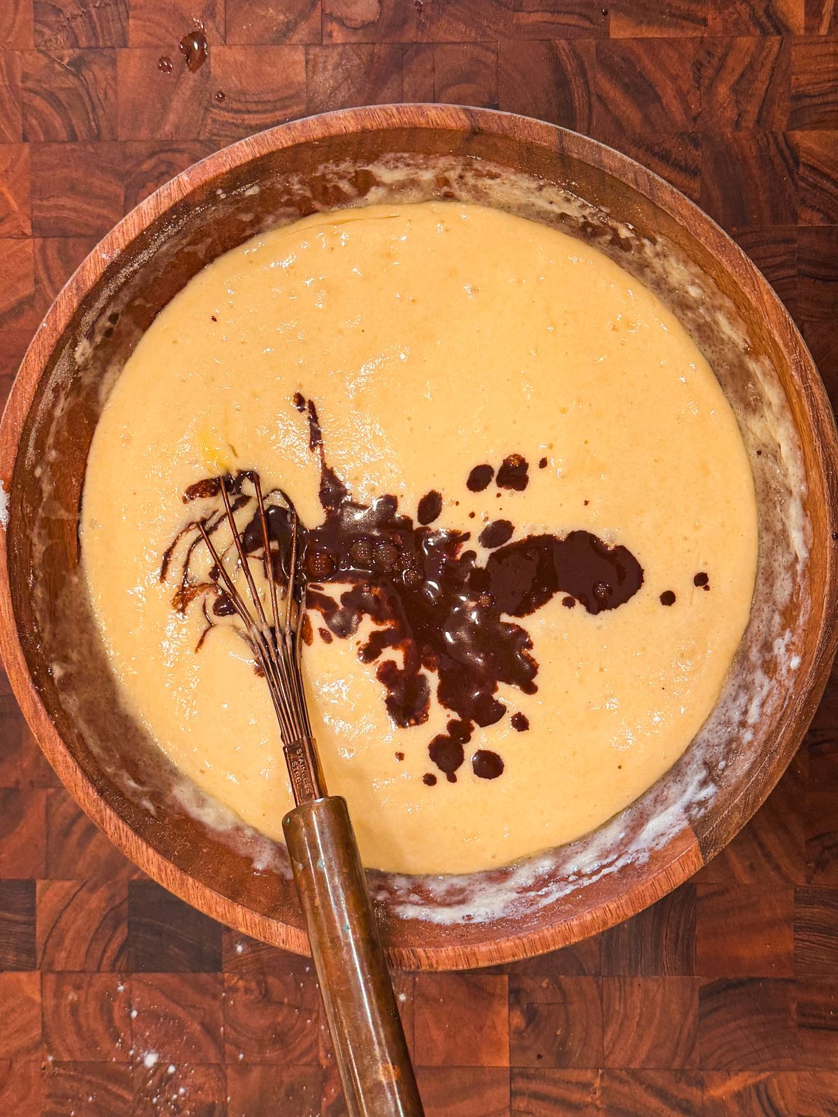 A wooden bowl filled with yellow cake batter, partially mixed with a small amount of melted chocolate and a metal whisk resting inside, on a wooden surface.