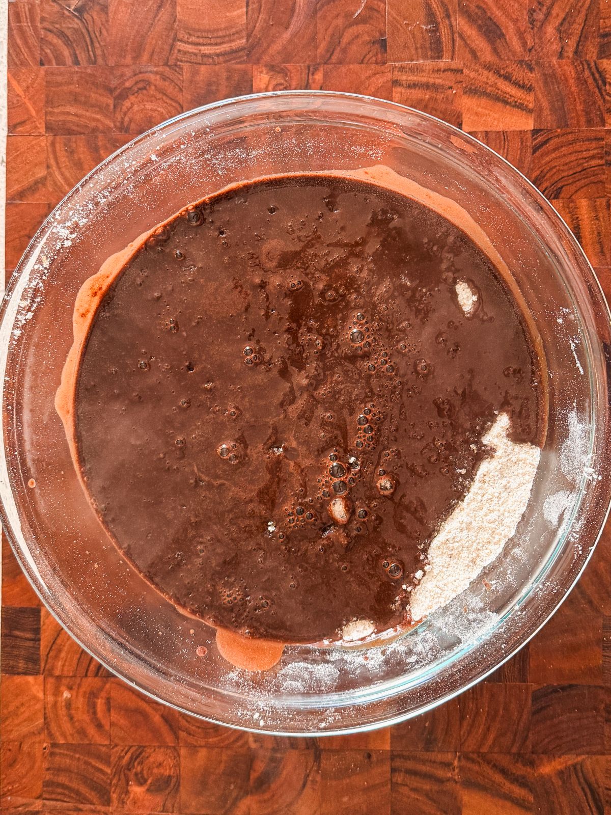 A glass bowl sits on a wooden surface, filled with a mixture of dark chocolatey liquid and visible patches of white flour, ready to be blended for baking.