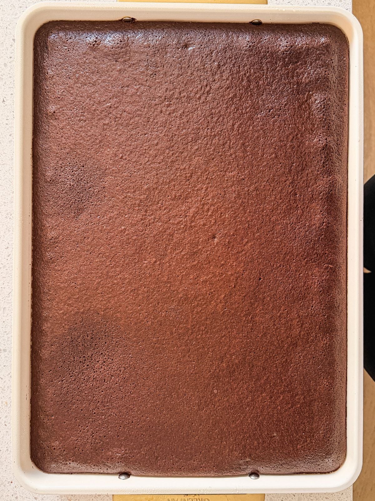A rectangular baking pan filled with a freshly baked, uncut chocolate cake with a smooth, slightly glossy surface.