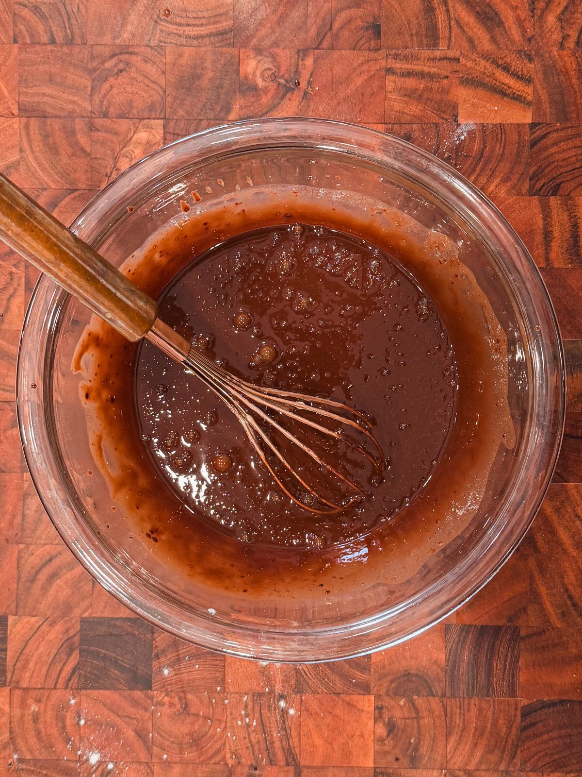 A glass bowl filled with chocolate batter being mixed with a whisk, placed on a wooden countertop.