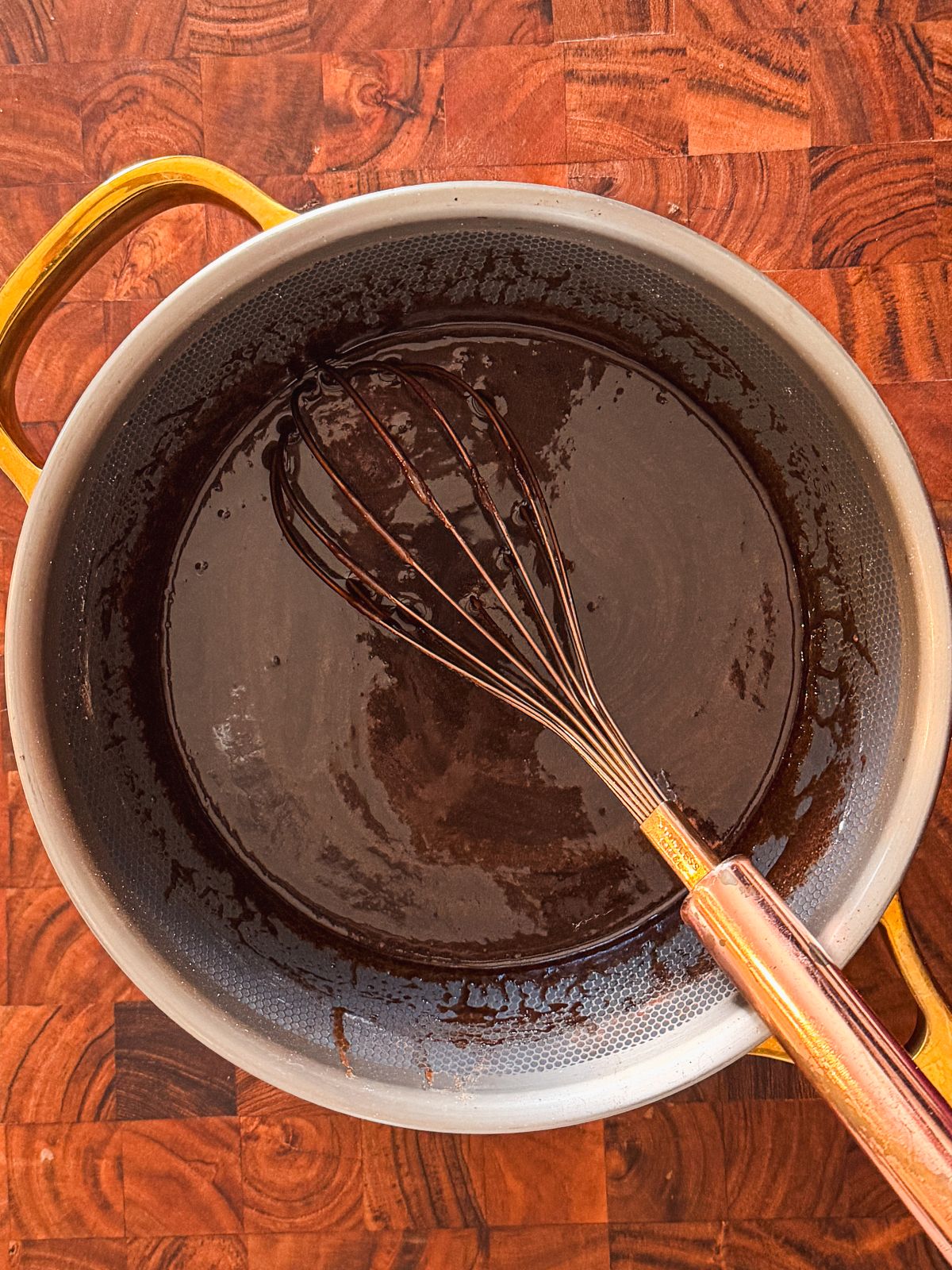 A metal whisk rests in a saucepan filled with glossy, dark chocolate mixture on a wooden countertop. The pan has yellow handles and textured interior.