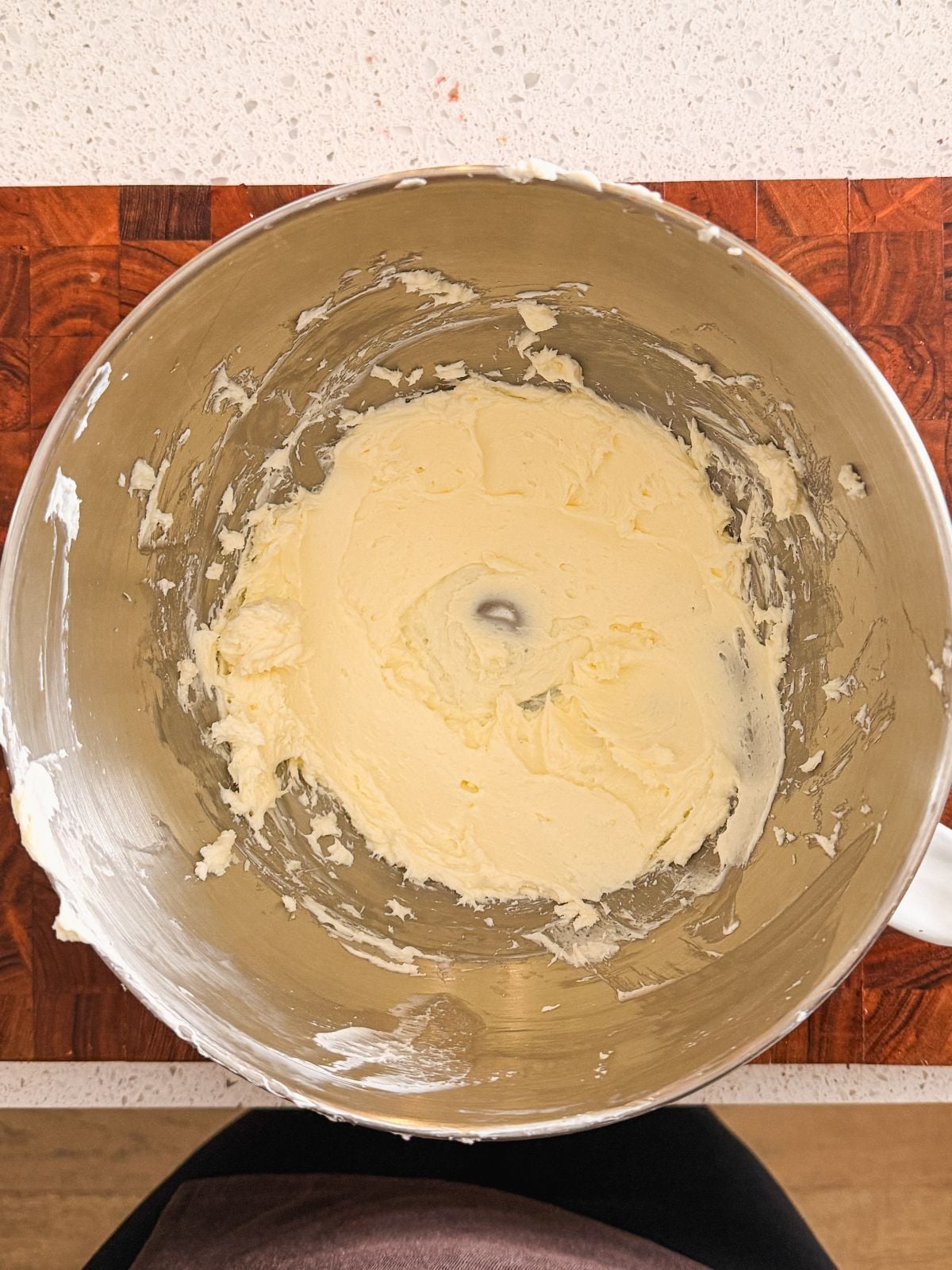 A mixing bowl containing cream cheese and sugar sits on a wooden countertop, viewed from above. The mixture is smooth and pale yellow, clinging to the sides of the bowl.