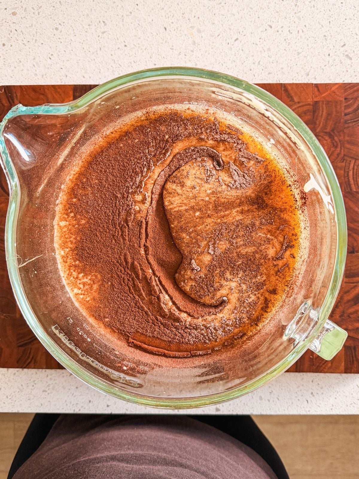 A glass mixing bowl filled with whipped eggs and sugar and sifted dry ingredients on top on a wooden cutting board, seen from above on a light countertop.
