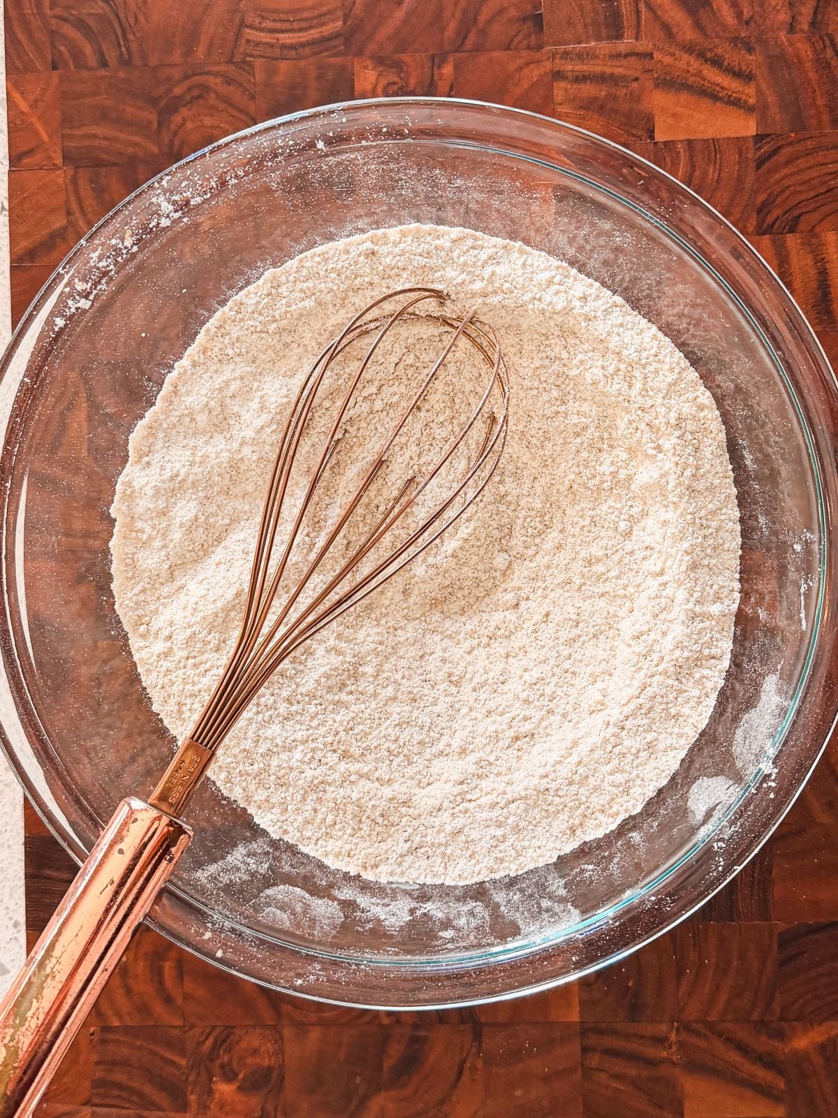 A glass bowl filled with flour sits on a wooden surface, with a metal whisk resting inside the bowl.