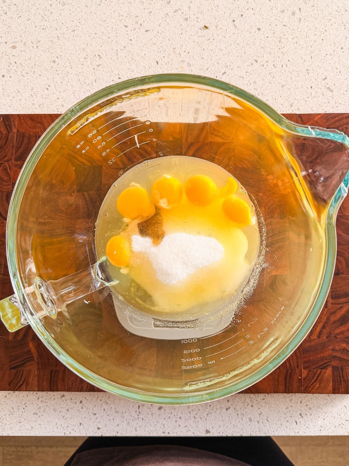 A glass mixing bowl containing six cracked eggs, granulated sugar, and vanilla extract sits on a wooden surface, viewed from above.