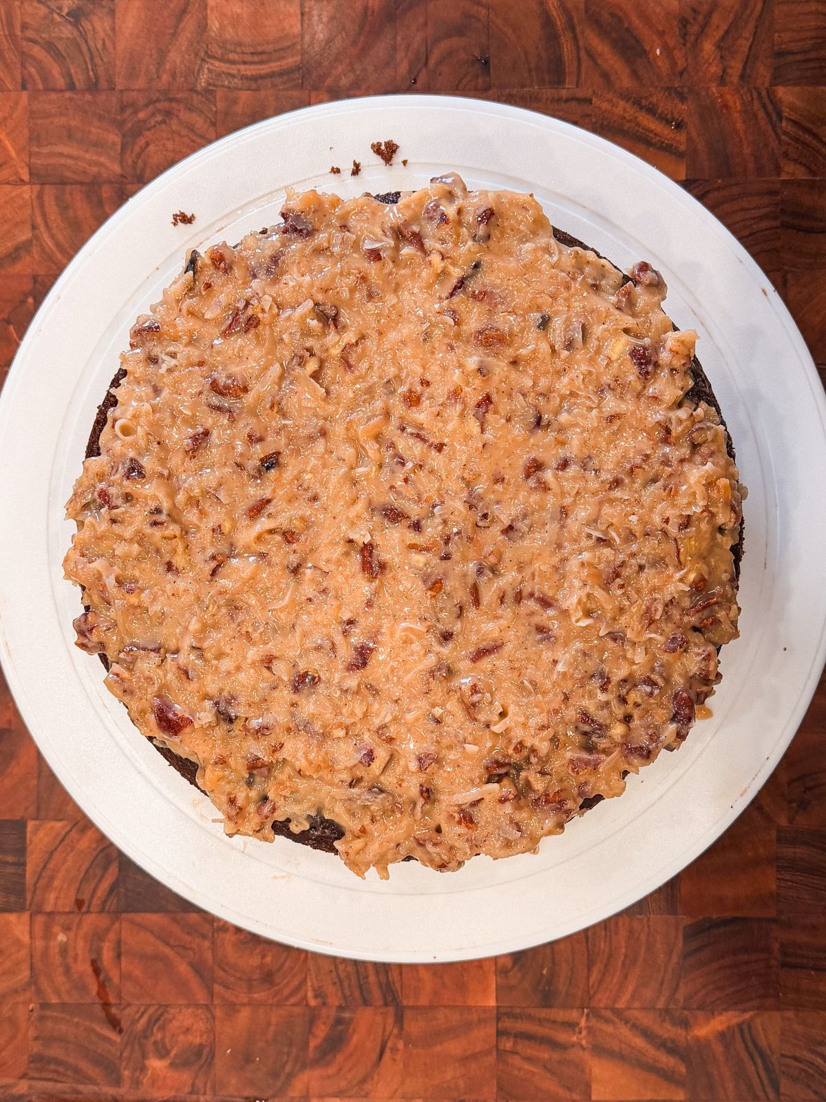 A round German chocolate cake topped with a thick layer of coconut-pecan frosting sits on a white plate atop a wooden surface.