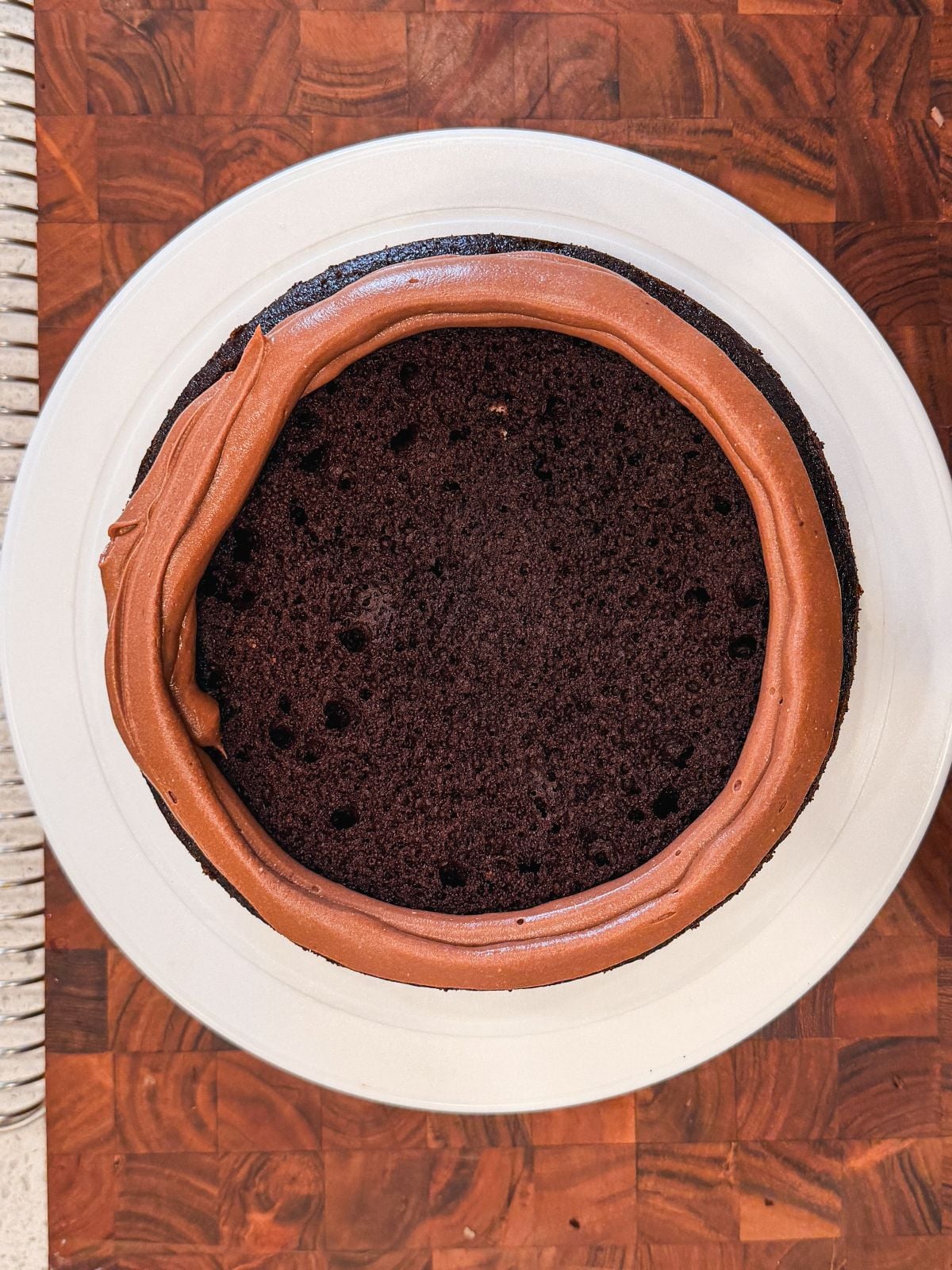 A round chocolate cake on a white plate, with a ring of chocolate frosting piped around the edge, sitting on a wooden surface. The cake's center is unfrosted and shows a rich, dark chocolate crumb.