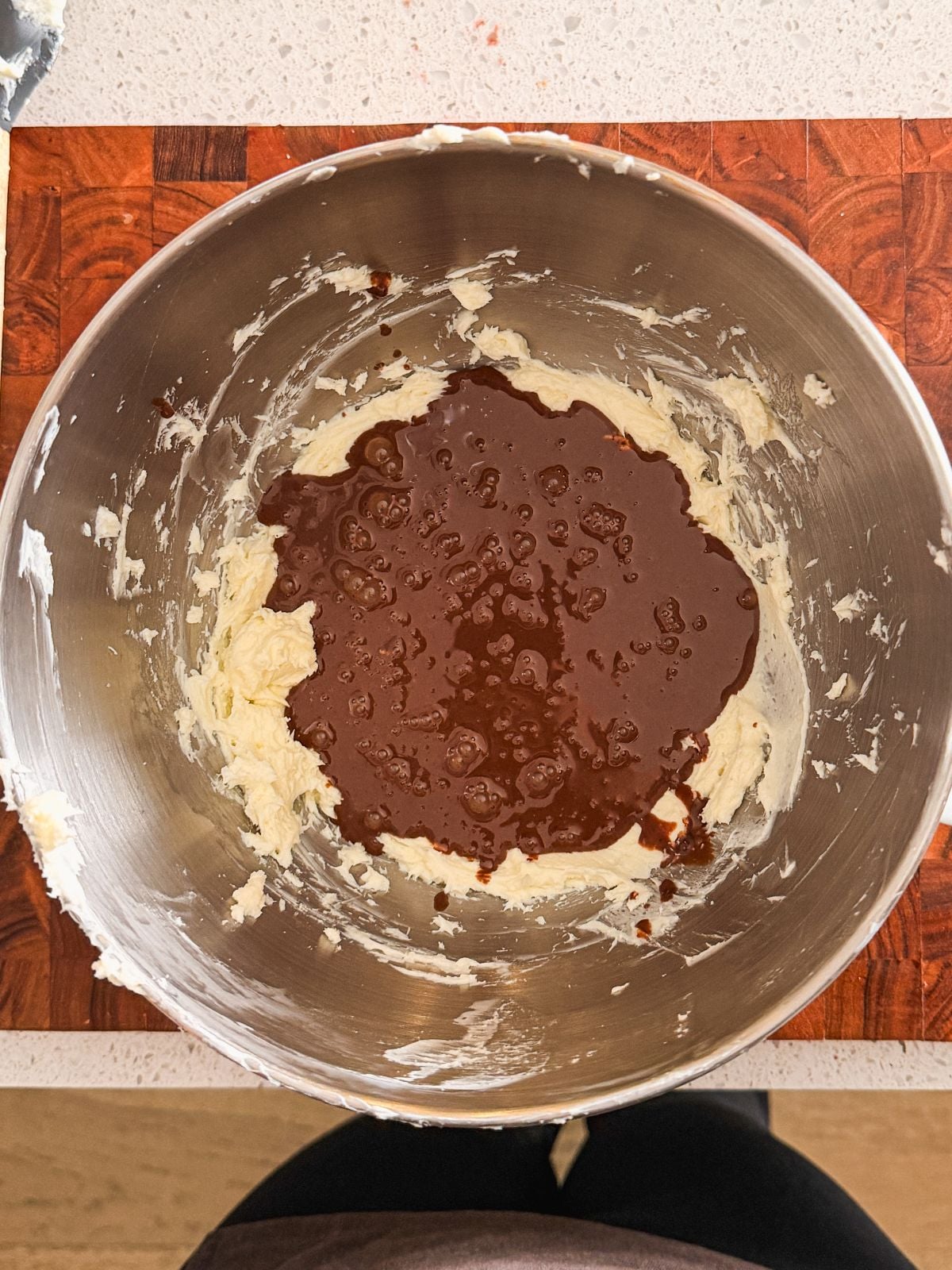 A stainless steel mixing bowl containing cream cheese and sugar, with melted chocolate poured on top, on a wooden cutting board.