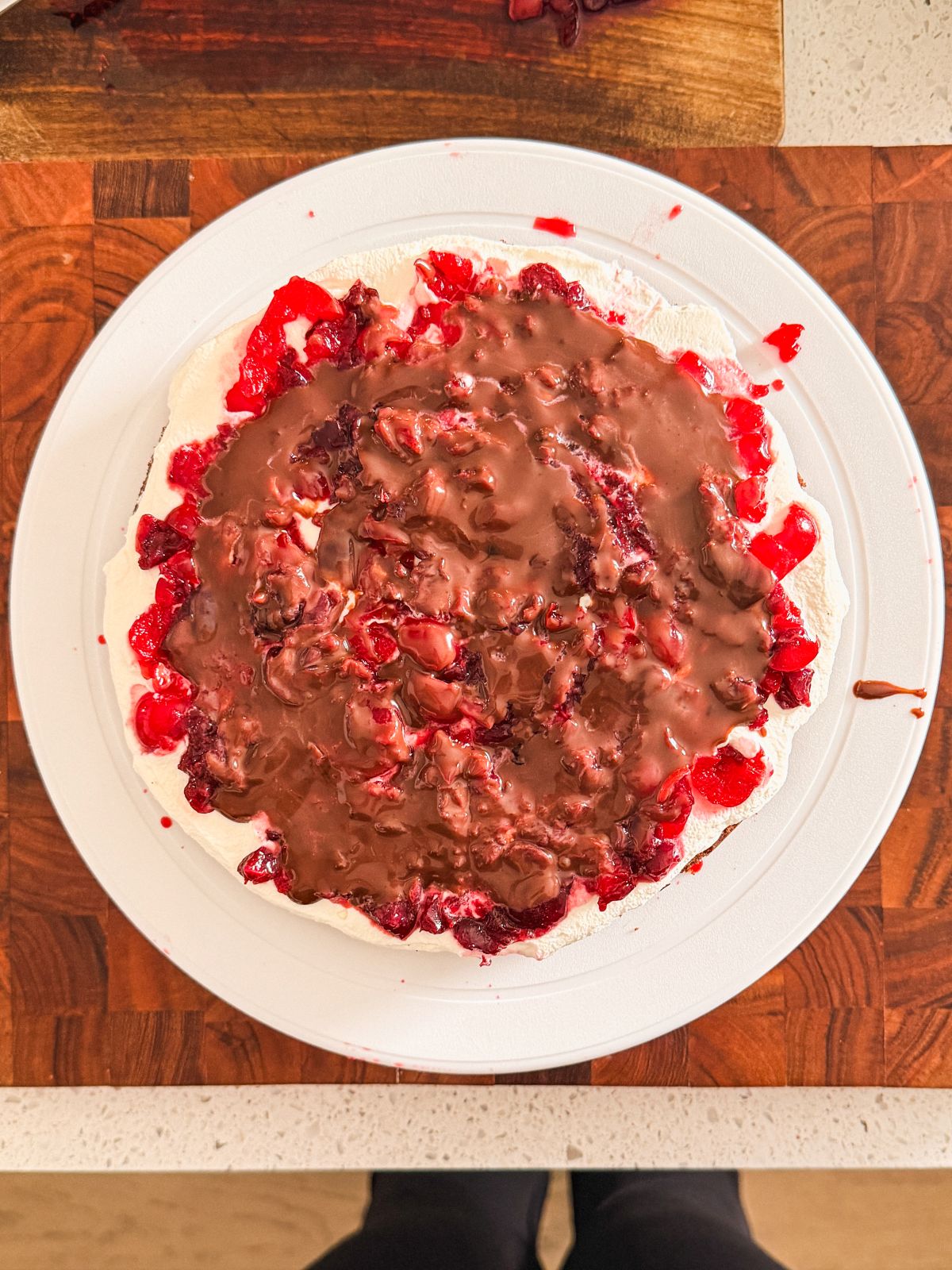 A round cake on a white plate features a chocolate cake covered with a layer of whipped cream, topped with chopped red fruit and a drizzle of ganache, sitting on a wooden cutting board.