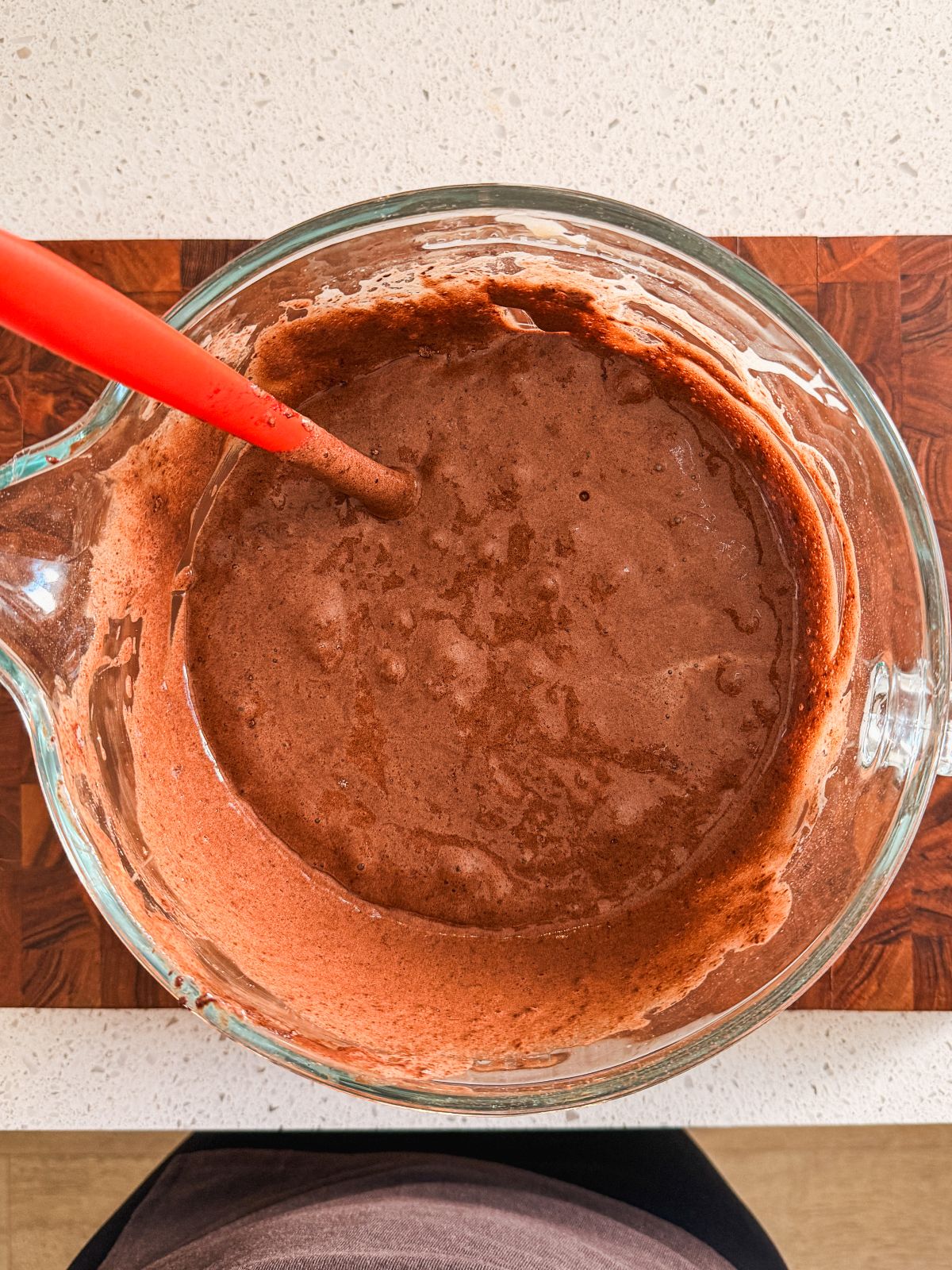 A glass mixing bowl filled with chocolate cake batter sits on a wooden cutting board, with a red spatula resting inside the bowl.