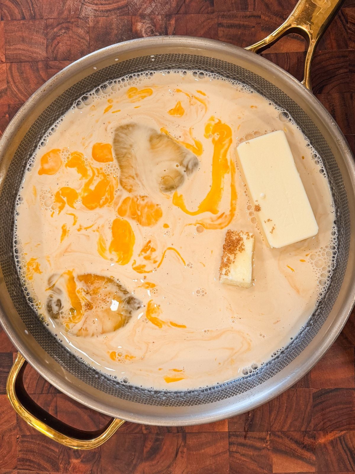 A metal pan on a wooden surface contains a creamy brown liquid with swirls of yellow oil, two chunks of butter