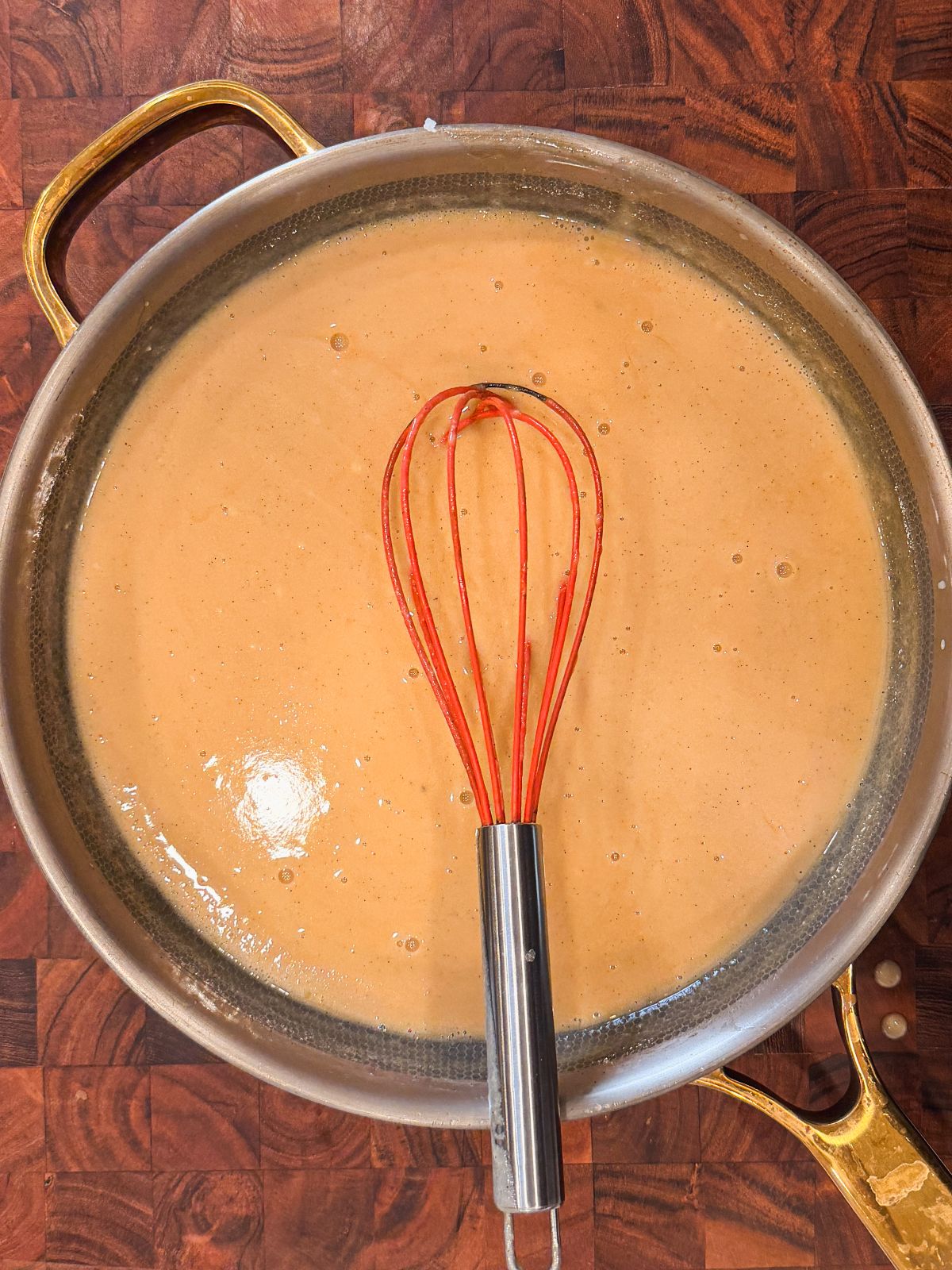 A saucepan filled with creamy, light brown butterscotch sauce sits on a wooden surface. A red-handled whisk rests in the sauce, indicating it has just been stirred or mixed.