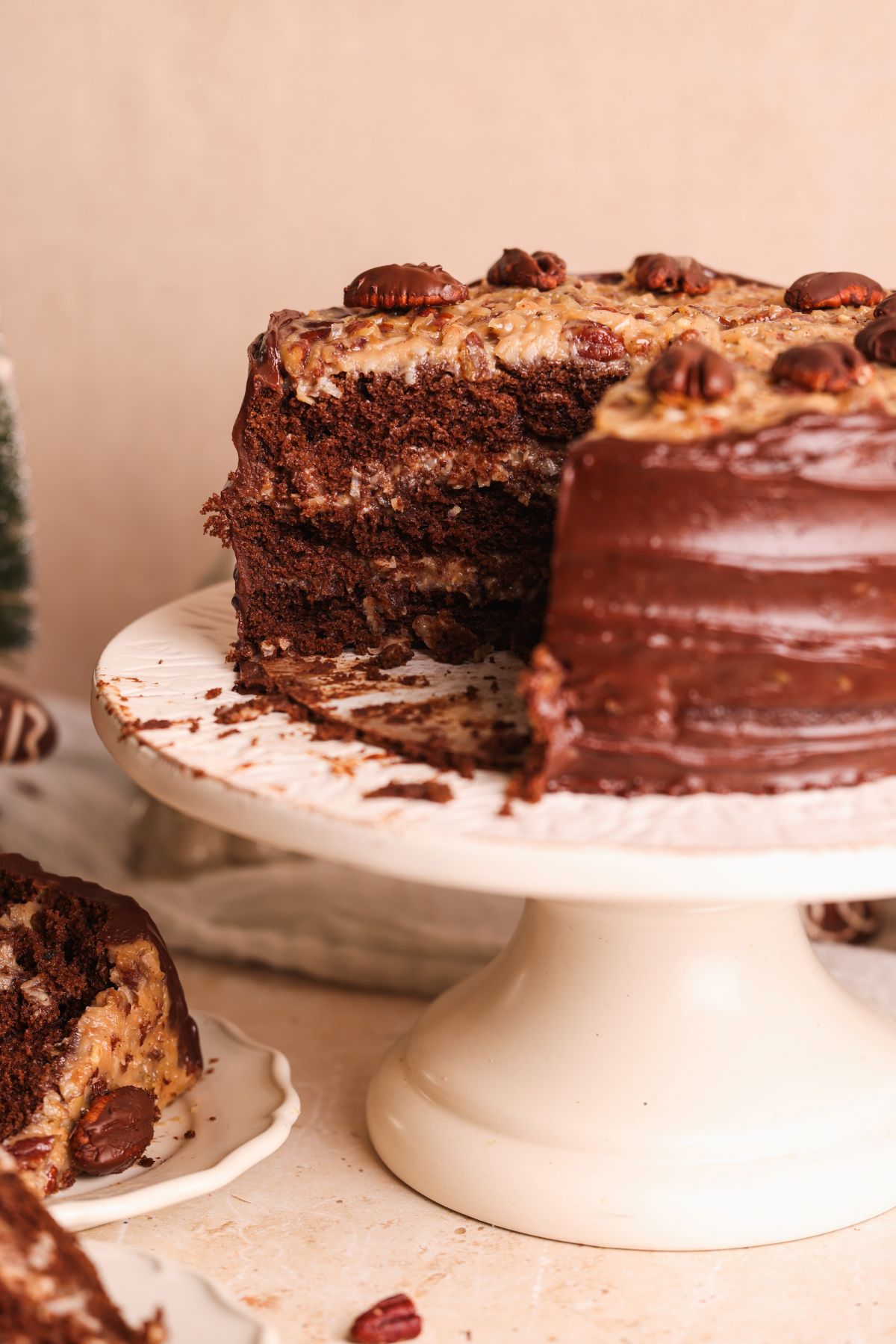 A german chocolate layer cake with a slice removed, topped with pecans and coconut-pecan frosting, displayed on a white cake stand. A slice of cake is on a separate plate nearby.