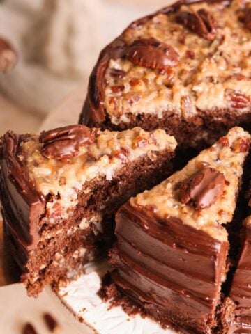 A slice of German chocolate cake being lifted from a whole cake. The cake has rich chocolate layers, chocolate frosting on the sides, and a coconut-pecan topping with pecan halves on top.