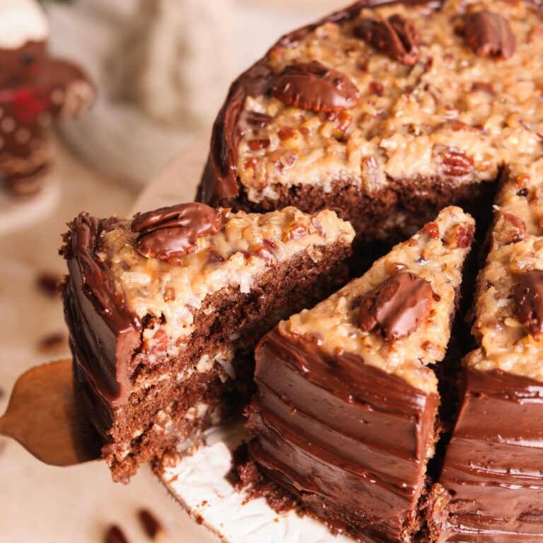 A slice of German chocolate cake being lifted from a whole cake. The cake has rich chocolate layers, chocolate frosting on the sides, and a coconut-pecan topping with pecan halves on top.