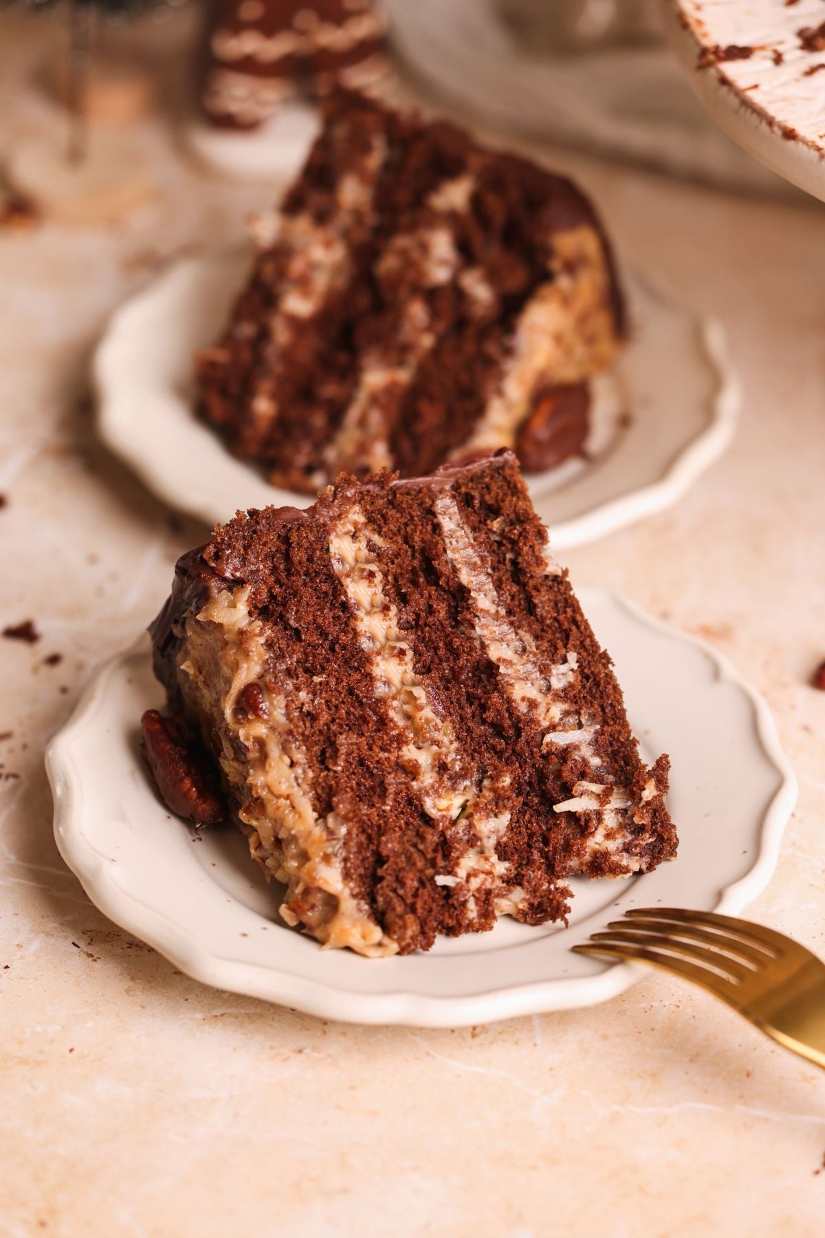 Two slices of layered german chocolate cake with creamy filling are served on white plates. A golden fork rests beside one slice, and the background is a light-colored surface with some crumbs scattered around.