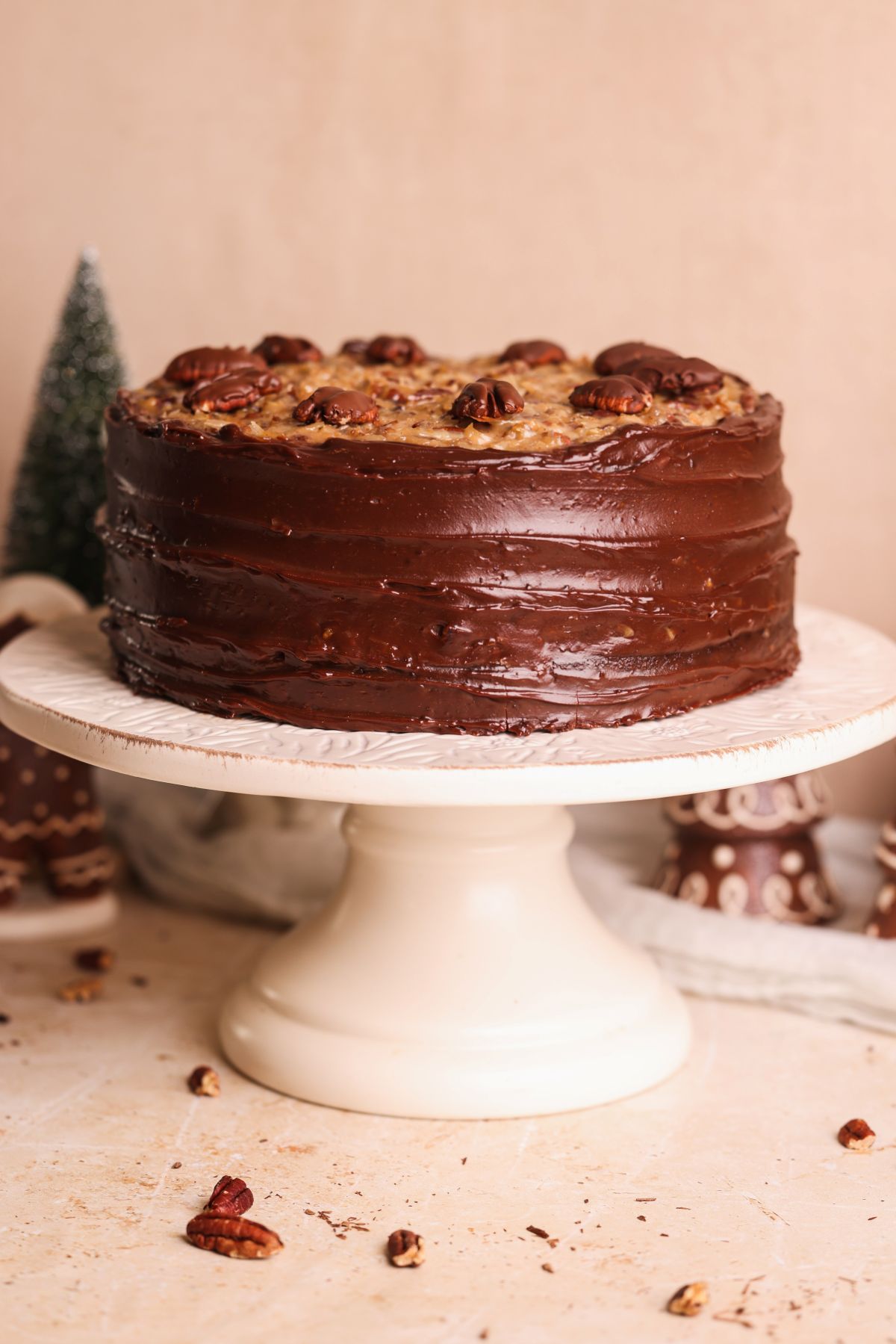 A German chocolate cake with chocolate frosting and pecan topping sits on a white cake stand. Decorated with whole pecans, the cake is displayed in a festive setting with blurred holiday decor in the background.