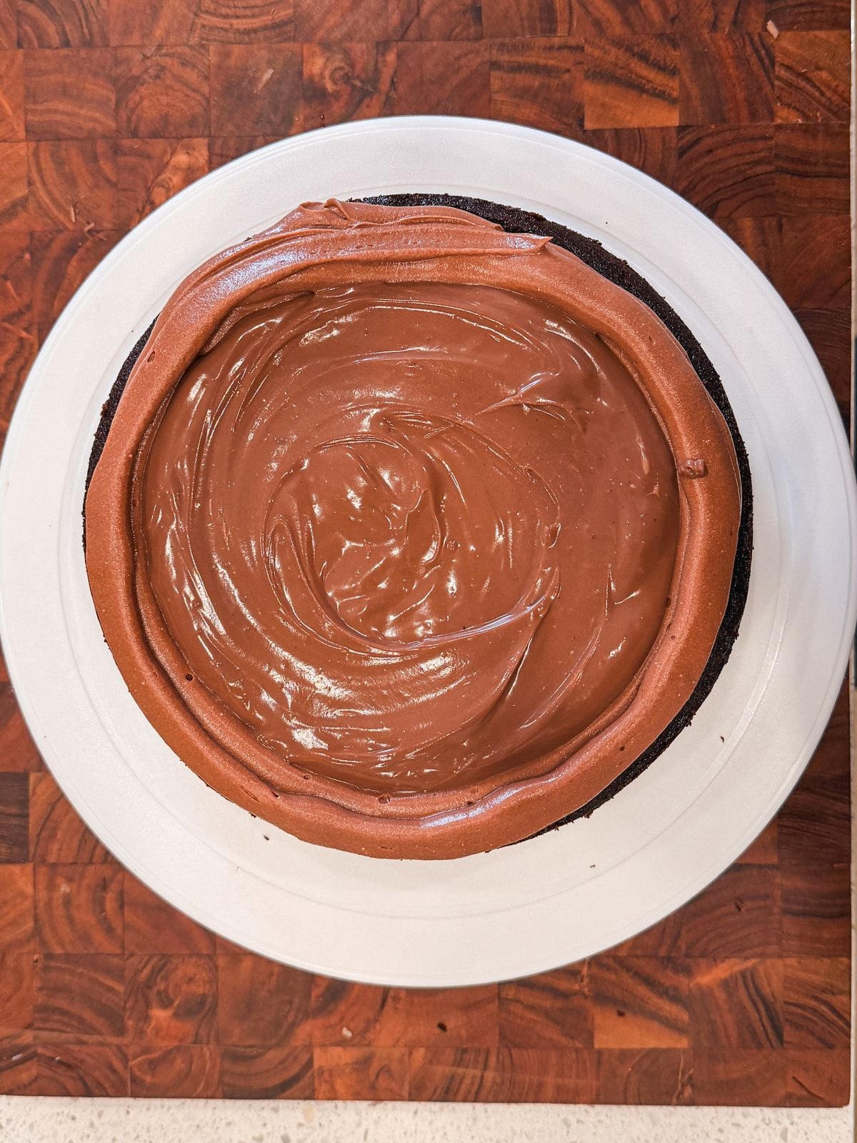 A round chocolate cake with a smooth, glossy chocolate filling and a dark crust, sitting on a white plate atop a wooden surface.