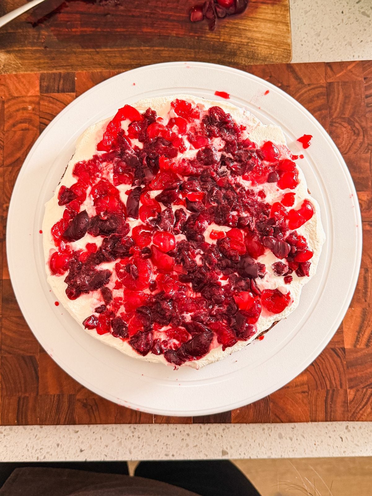 A round cake topped with a mixture of chopped red and dark cherries sits on a white plate, placed on a wooden cutting board. The cake has a creamy white layer beneath the fruit.