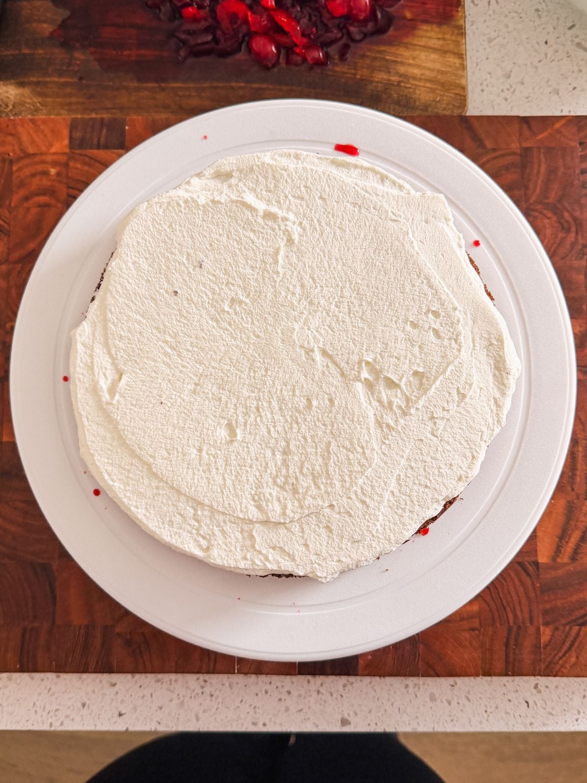 A round cake covered with a layer of white frosting sits on a white plate, placed on a wooden surface. Red crumbs or pieces are visible around the edge and on a nearby cutting board in the background.