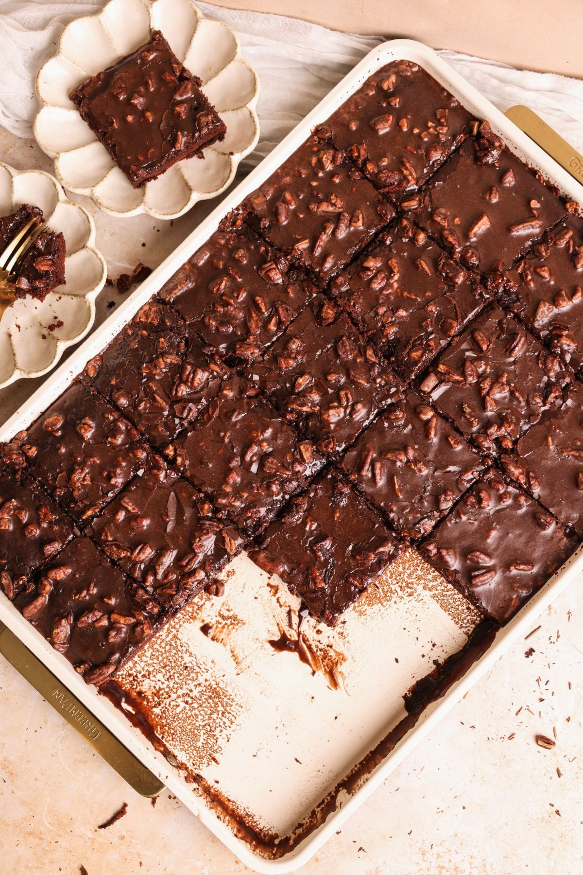 A tray of texas sheet cake, most cut into squares with a few pieces missing. One slice is placed on a small white plate beside the tray, along with some crumbs and a fork.