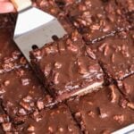 A metal spatula lifts a texas sheet cake slice topped with chopped pecans from a tray of cake, all covered in glossy chocolate icing.