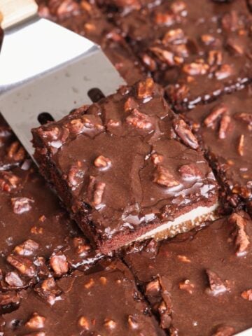 A metal spatula lifts a texas sheet cake slice topped with chopped pecans from a tray of cake, all covered in glossy chocolate icing.
