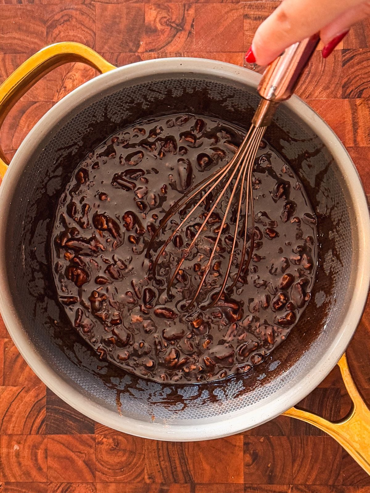 A hand holding a whisk stirs a glossy, dark chocolate mixture with nuts in a saucepan with gold handles, placed on a wooden surface.