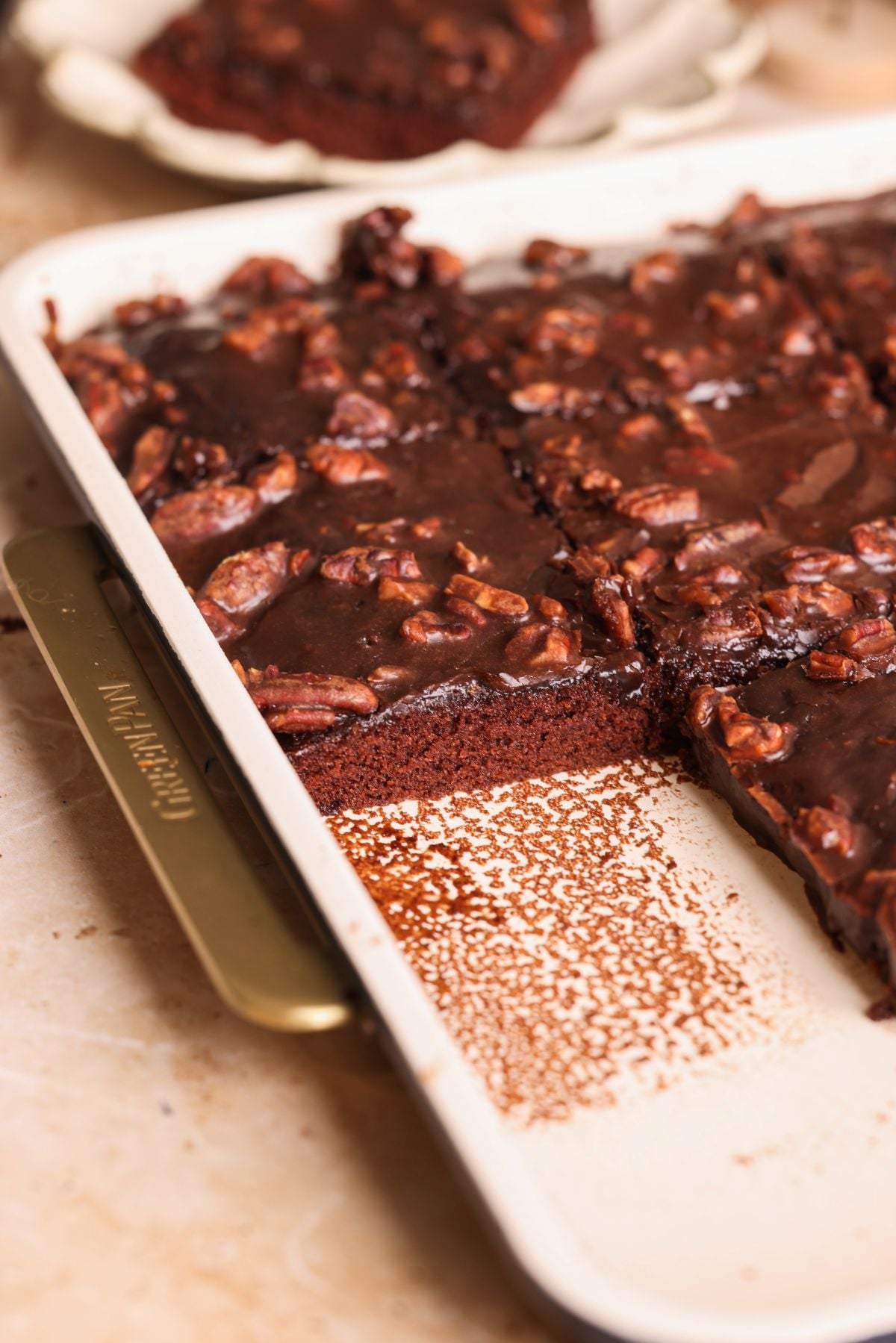 A baked chocolate sheet cake topped with chopped nuts and chocolate glaze sits in a rectangular pan with a few pieces cut out, revealing the moist cake and a clean edge where slices have been removed.