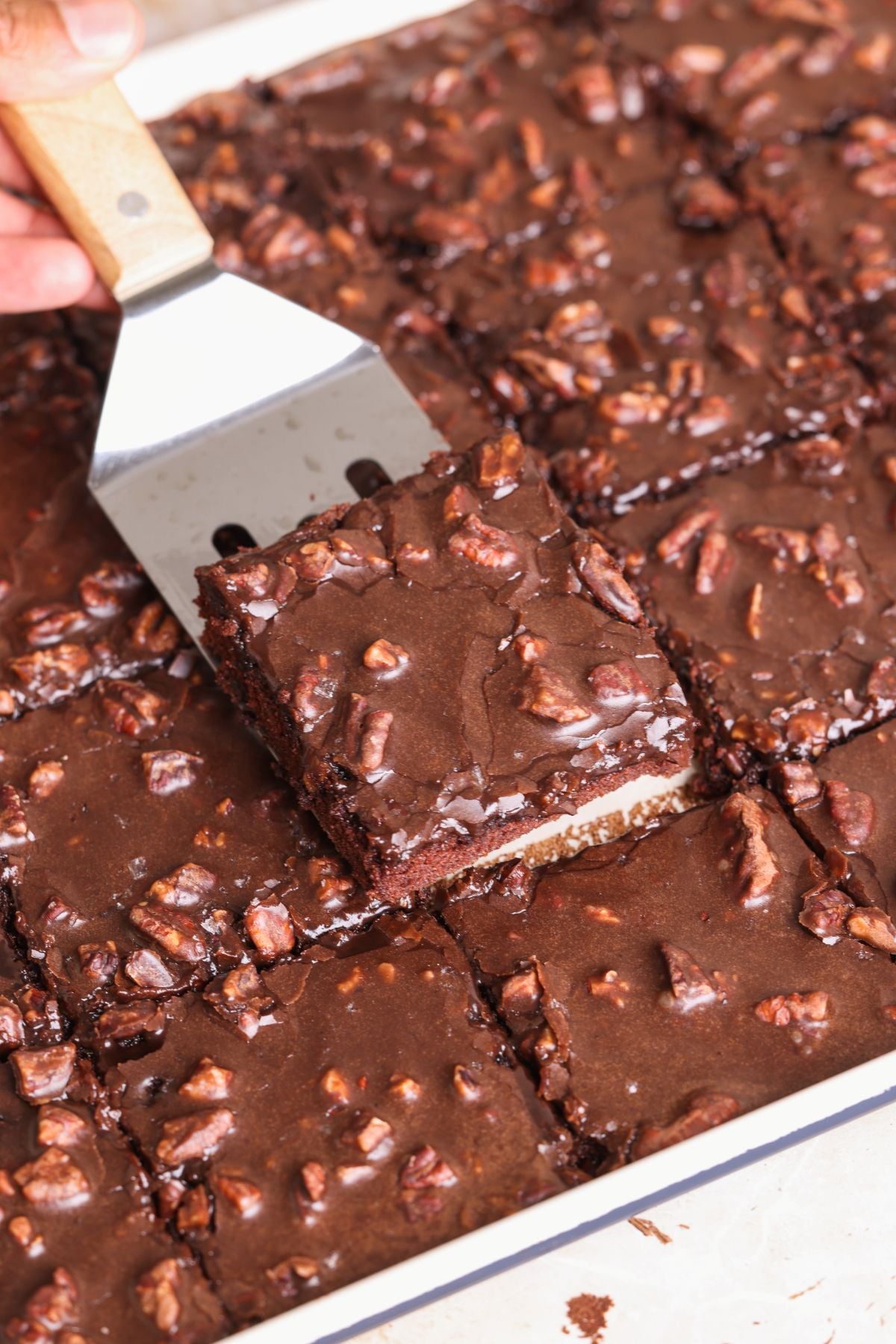 A spatula lifts a square slice of texas sheet cake with chopped nuts from a tray, all cut into even squares. The cake has a glossy, rich chocolate topping and visible nut pieces.