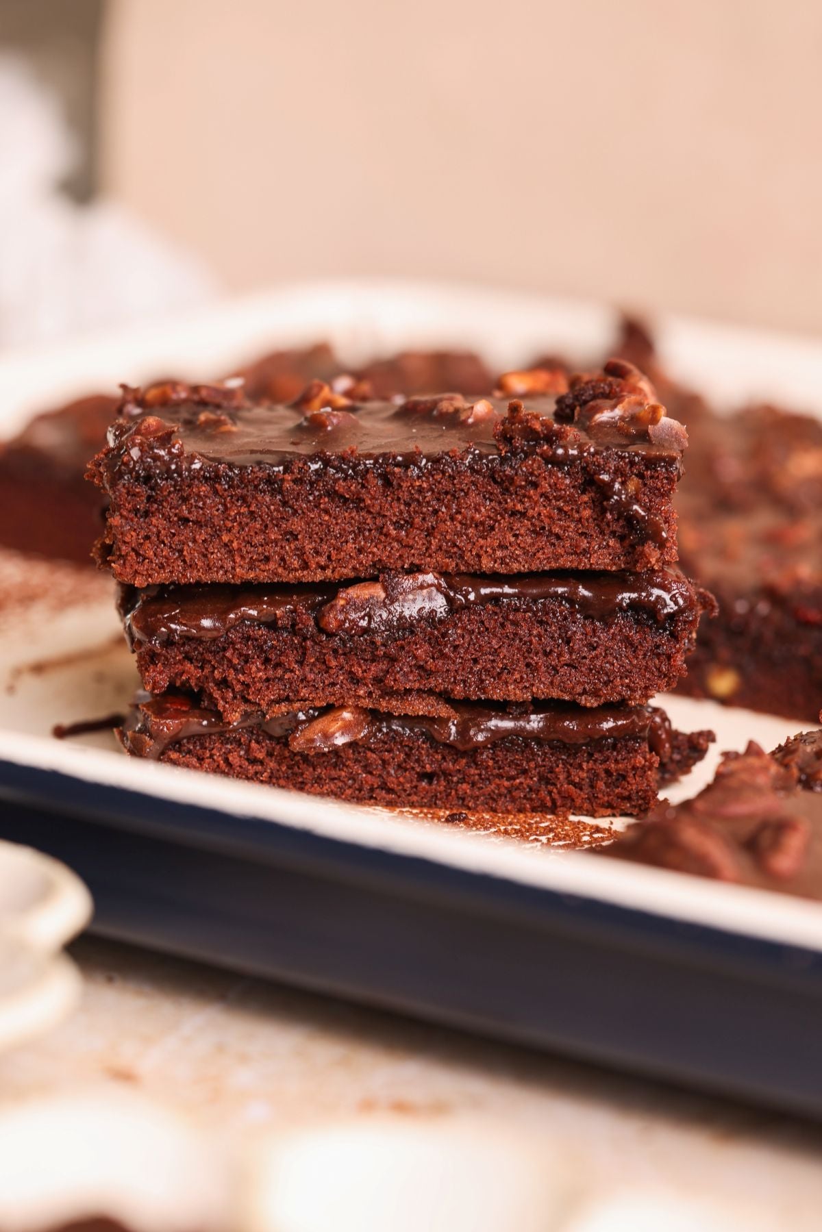 Three slices of texas sheet cake with a glossy chocolate and nut topping are stacked on a white tray. More slices are visible in the background.