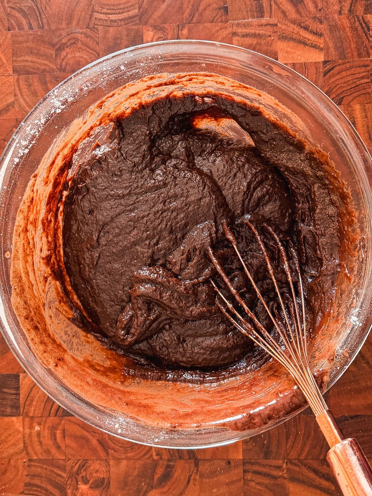 A glass bowl filled with thick chocolate batter sits on a wooden countertop. A metal whisk, partially covered in the batter, rests inside the bowl.