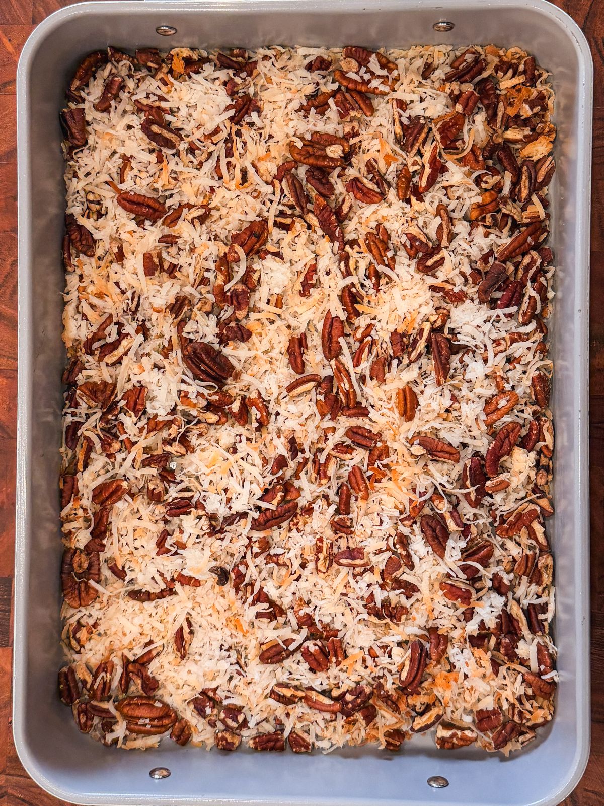 A rectangular metal baking pan filled with shredded coconut and chopped pecans, evenly spread across the surface.