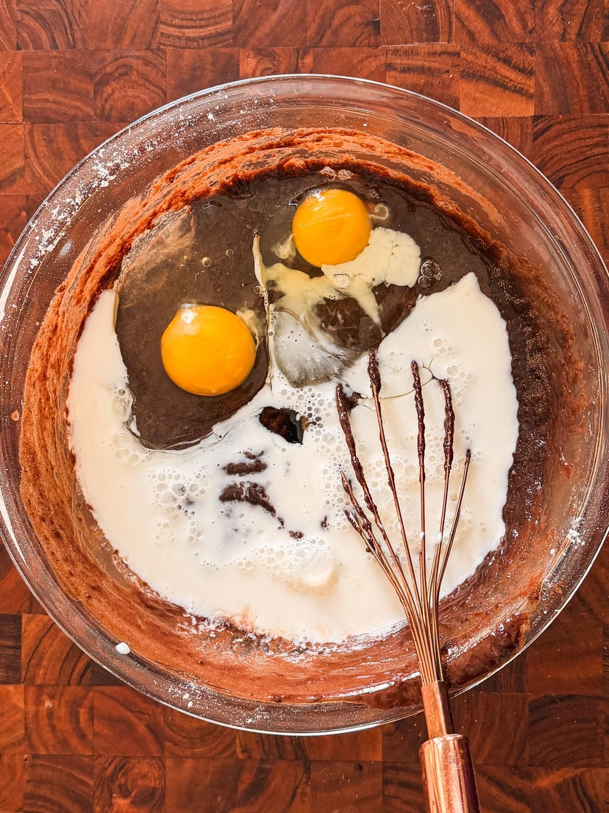 A glass bowl contains a chocolate batter mixture with two raw eggs, milk, and flour. A metal whisk is resting inside the bowl, ready to mix the ingredients, all placed on a wooden surface.