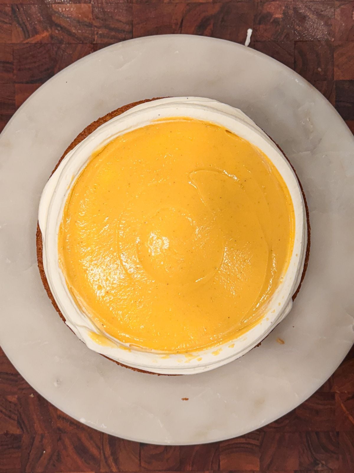 A round cake on a white marble platter, topped with a smooth, bright yellow layer of mango curd and a white ring of frosting around the edge, viewed from above on a dark wooden surface.