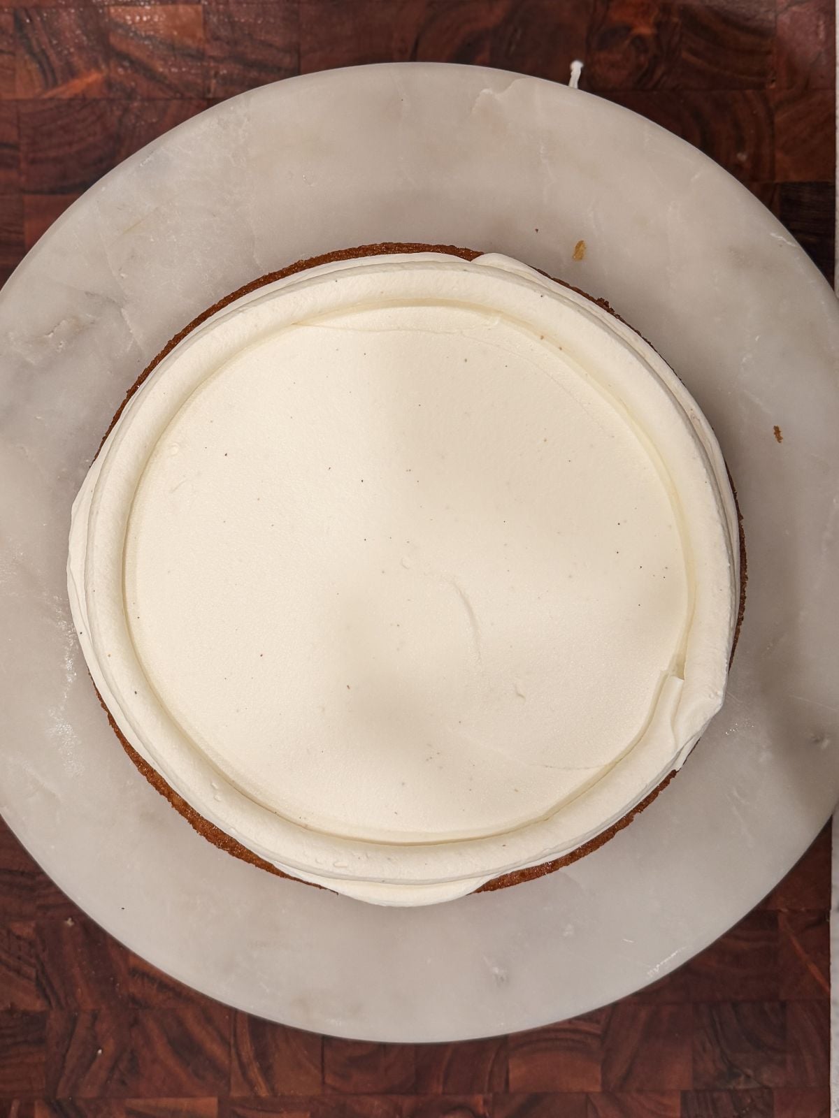 A round cake with smooth white frosting sits on a white marble plate, viewed from above, on a wooden surface.