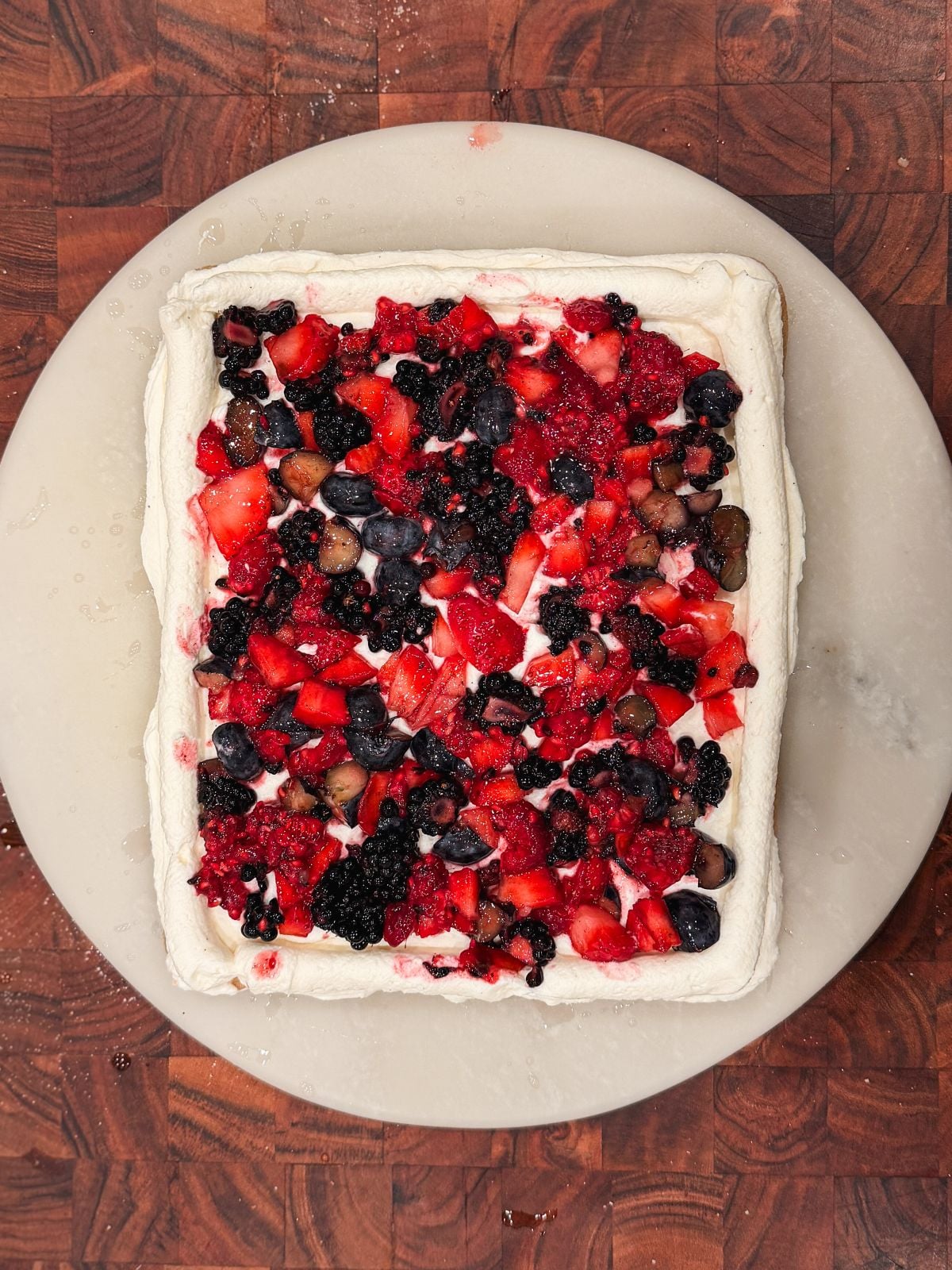A rectangular cake topped with whipped cream and a mix of strawberries, blueberries, and blackberries, sits on a round white plate over a wooden surface.