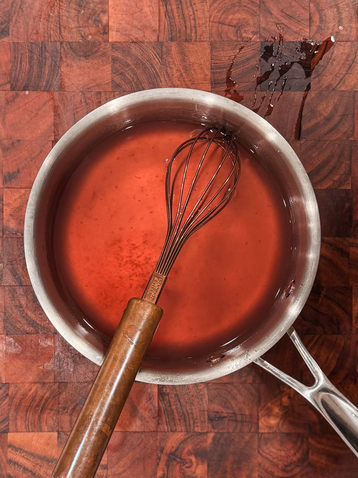 A saucepan filled with a red strawberry simple syrup and a whisk resting inside, placed on a wooden cutting board with a few drops of liquid spilled nearby.