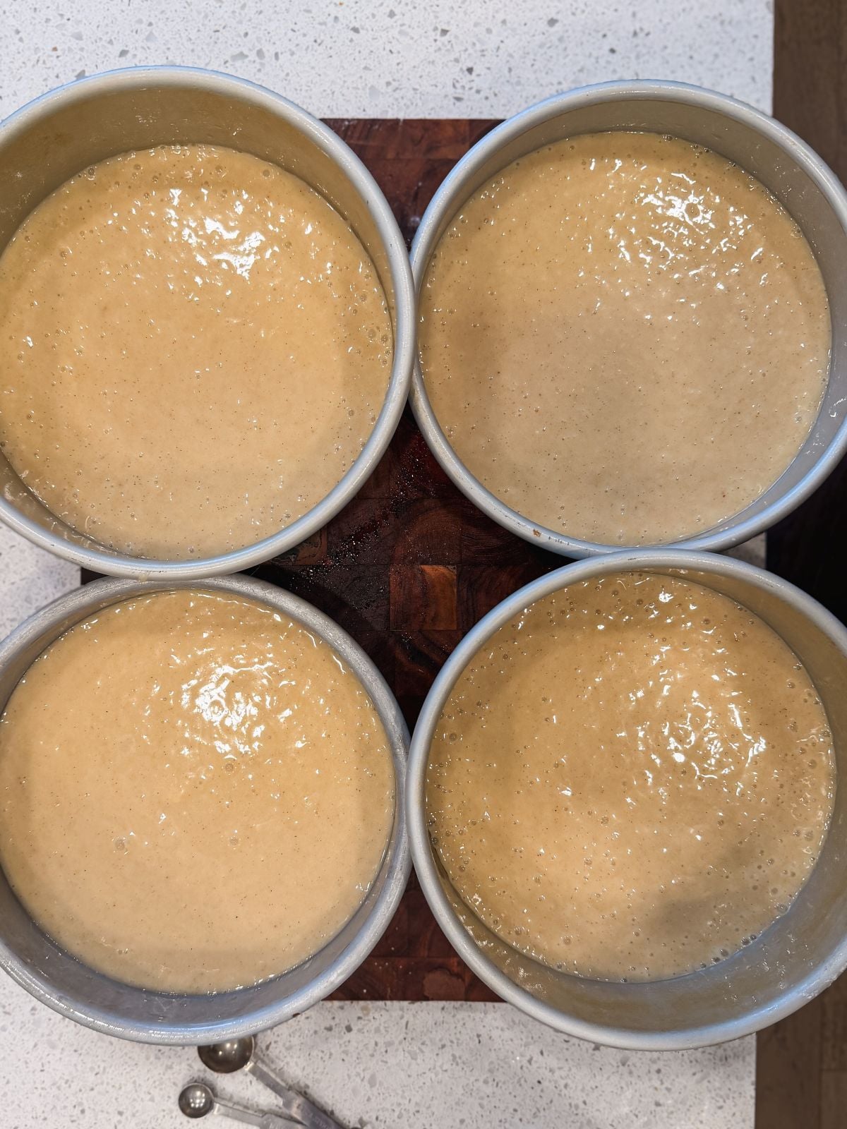 Four round cake pans filled with raw, light brown batter are arranged close together on a countertop, ready to be baked.