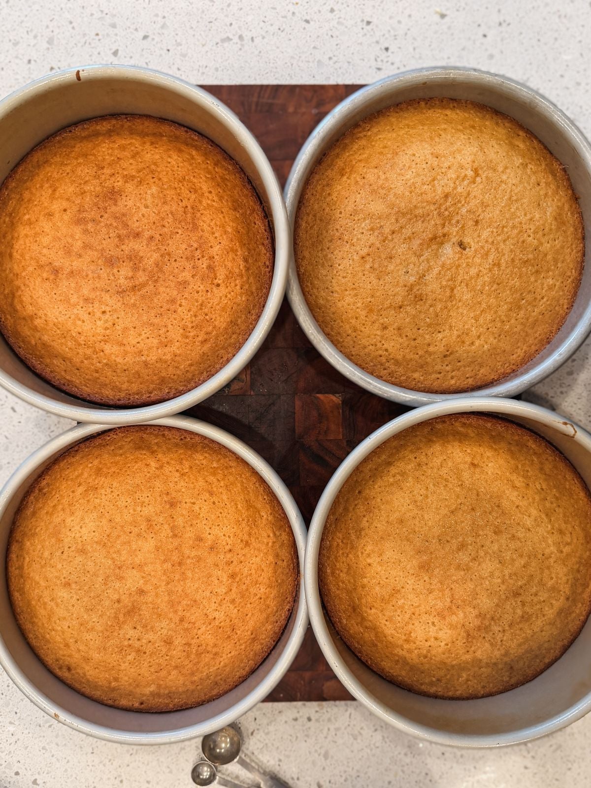 Four round, golden-brown cakes cooling in metal pans, arranged in a square on a wooden cutting board atop a light-colored countertop. A metal measuring spoon is partially visible in the bottom left corner.