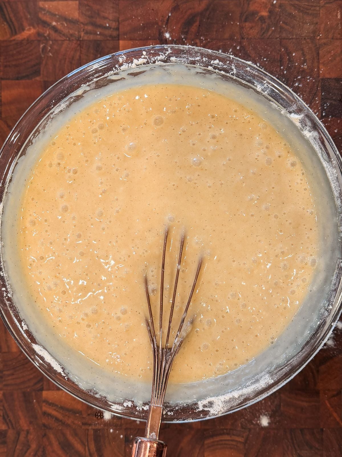 A glass bowl filled with cake batter sits on a wooden surface, with a metal whisk resting inside the bowl. Some flour is visible along the edges of the bowl.