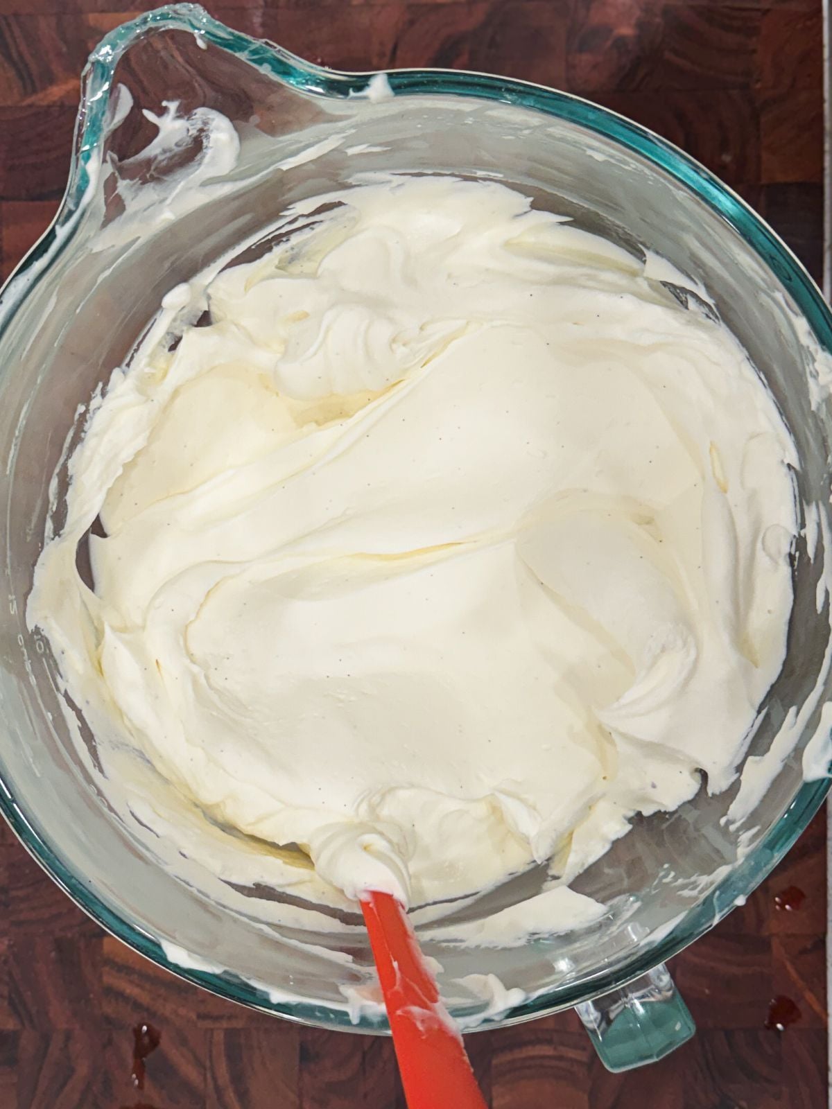 A glass mixing bowl filled with smooth, whipped cream cheese frosting. A red spatula is resting in the mixture, and the bowl is placed on a wooden surface.