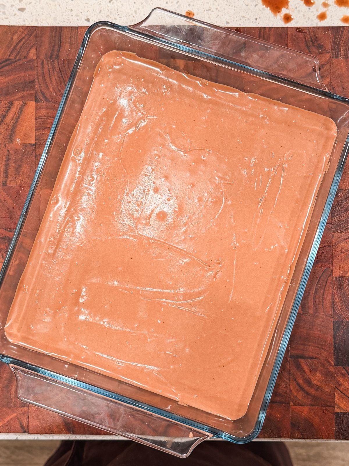 A glass baking dish filled with smooth, light brown batter sits on a wooden countertop, ready to be baked. Some splatters are visible on the counter in the background.